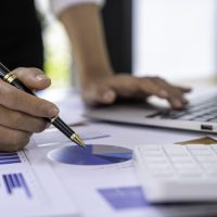 Businesswoman holding graph pen to analyze marketing plan, turnover and profit, calculating taxes with calculator and laptop on wooden desk in office.